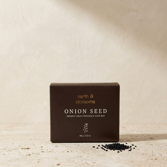 A dark brown rectangular box labeled 'earth & blossoms ONION SEED' stands on a textured beige surface with a small pile of black onion seed beads in front. The background is a warm, textured beige wall with soft shadows.