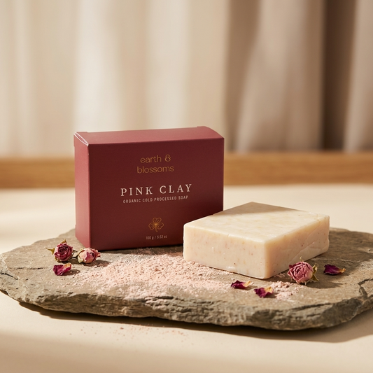 A bar of pale pink clay soap sits on a stone slab beside its dark red box labeled 'earth 8 blossoms PINK CLAY organic clay infused soap,' with scattered pink powder and dried rose buds in a softly lit setting.