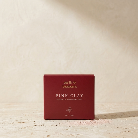 A square dark red box of earth & blossoms Pink Clay Organic Cold Processed Soap stands on a textured beige surface with a matching beige background.
