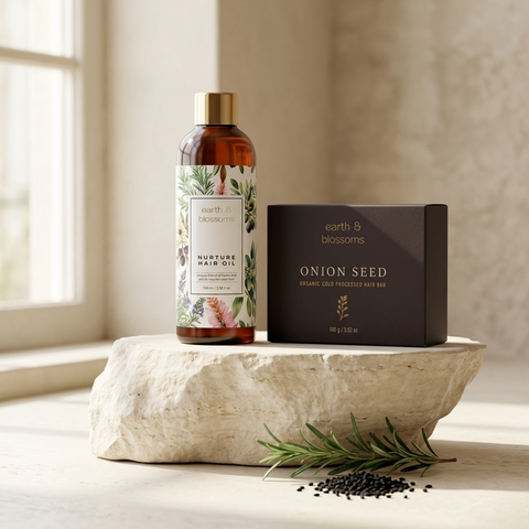 A bottle of earth & blossoms Nurture Hair Oil and a black box of earth & blossoms Onion Seed Organic Cold Proofed Hair Bar are styled on a round stone pedestal with loose seeds and rosemary sprigs in a warm, sunlit room.