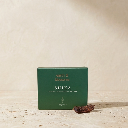 A dark green rectangular box of earth & blossoms SHIKA Organic Cold-Processed Hair Bar sits on a textured beige surface with a dried herbal ingredient resting in front of it.