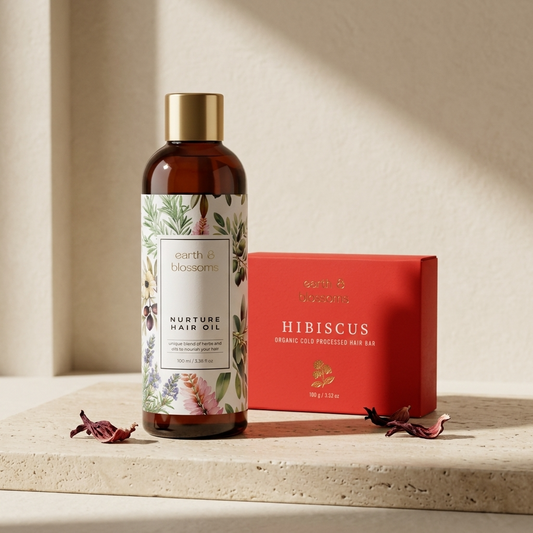 A bottle of earth & blossom Nurture Hair Oil with a gold cap stands next to a red gift box labeled Hibiscus, styled on a beige stone surface with dried hibiscus flowers and warm sunlight shadows.